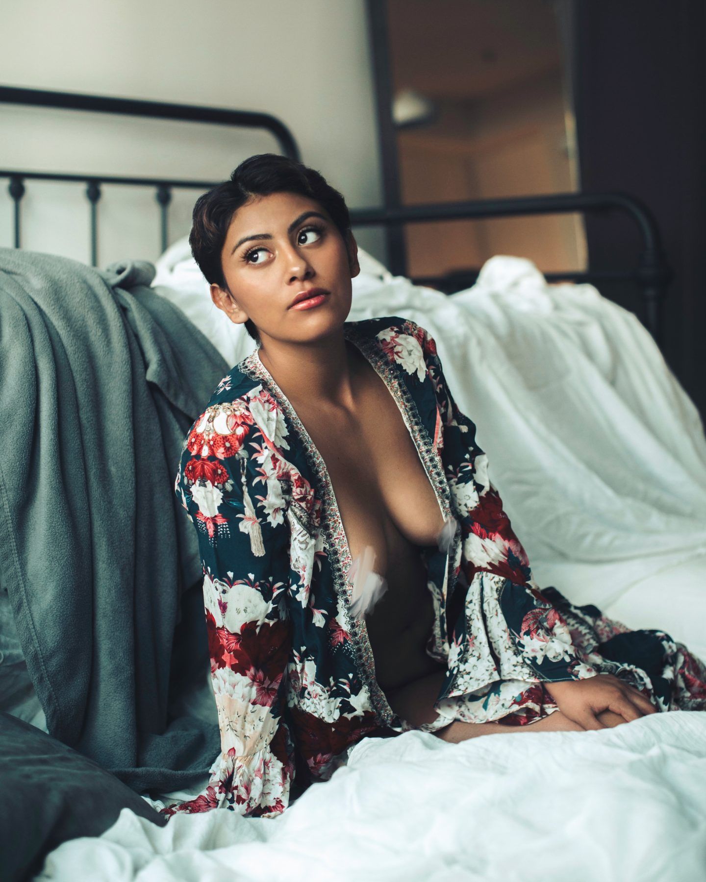 Woman in an open floral robe on a white bed beside an iron headboard